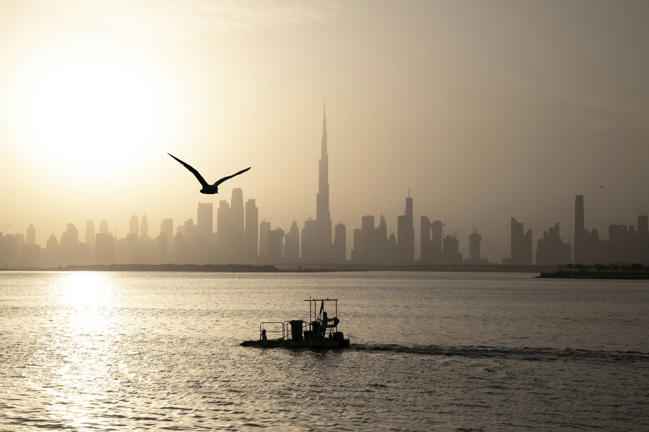 Hazy Dubai skyline with the Burj Khalifa across calm water, a gull in flight and a small work boat